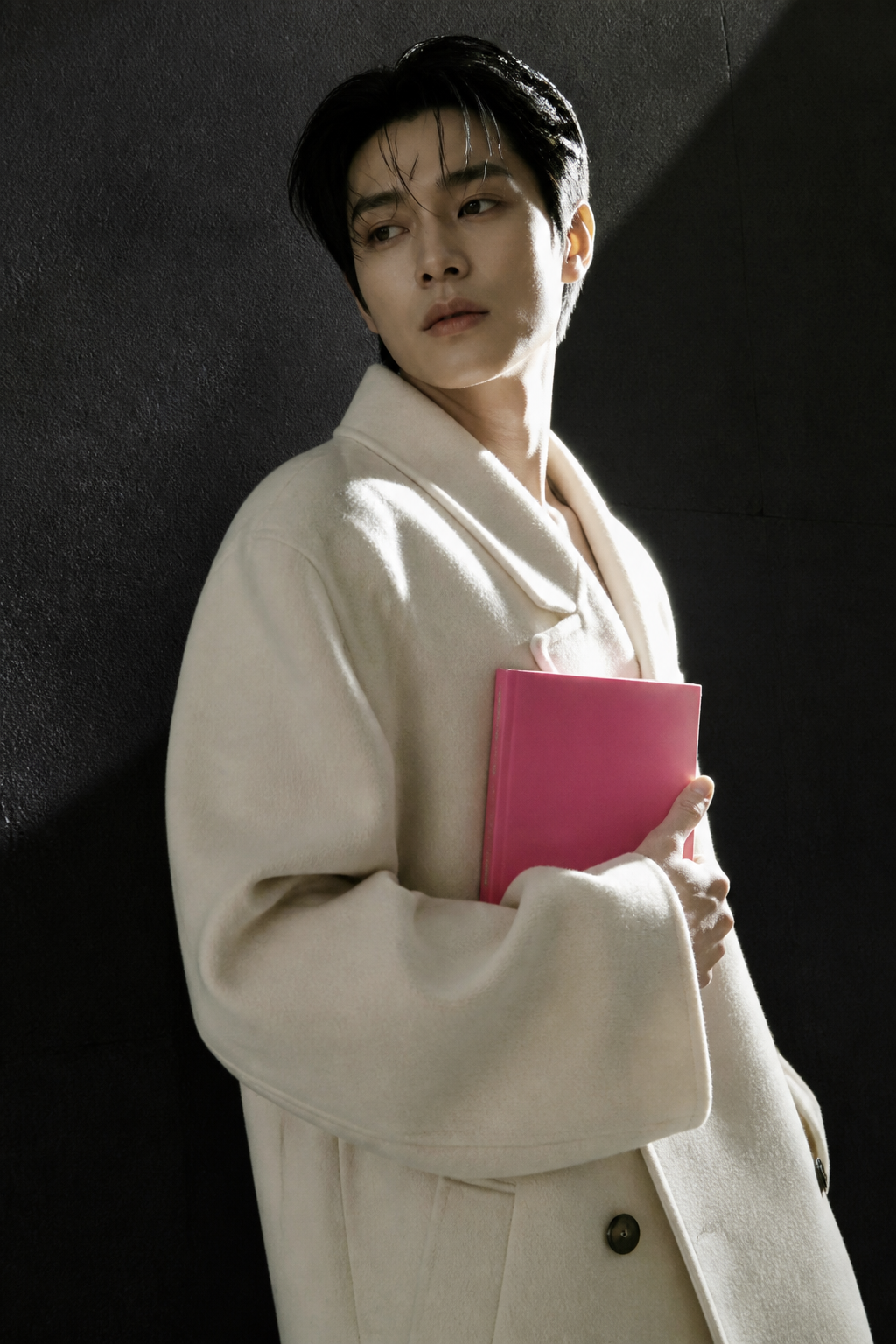 A person in a cream coat holding a pink notebook against a charcoal wall