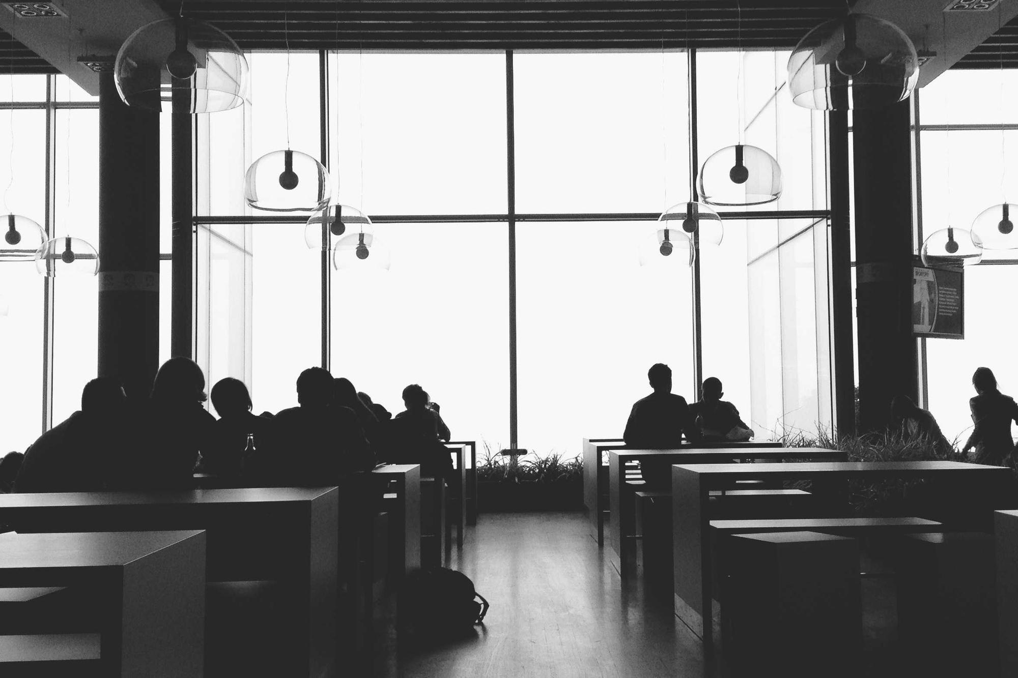 Silhouettes of people working at long tables in front of tall windows