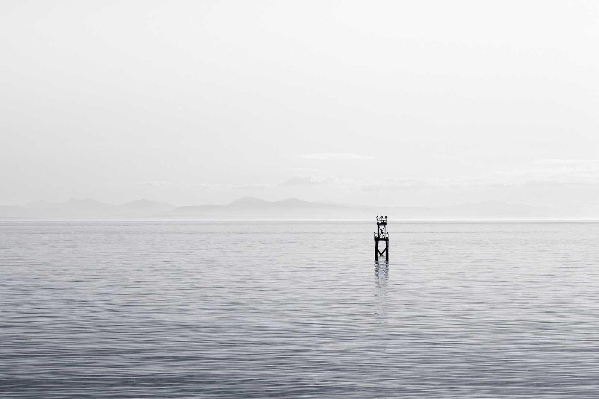 A still sea under a pale sky with a single marker post standing in the water
